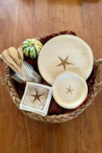 Wooden plates with star design, wooden utensils, and coasters in a woven basket on a wooden surface.