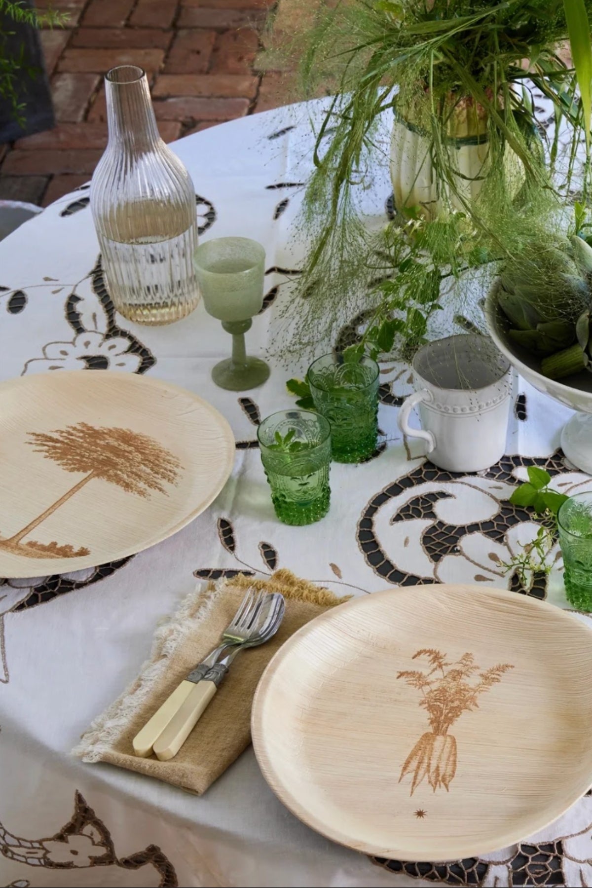 Garden party table setting with palm leaf plates, glasses, and cutlery on a decorative tablecloth.
