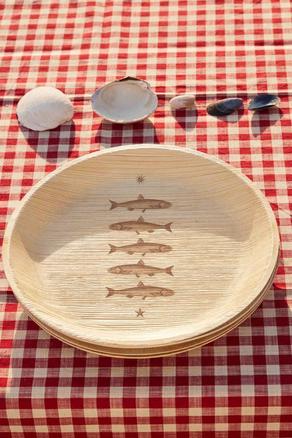Engraved compostable palm leaf plates with fish design styled on red gingham tablecloth with seashells