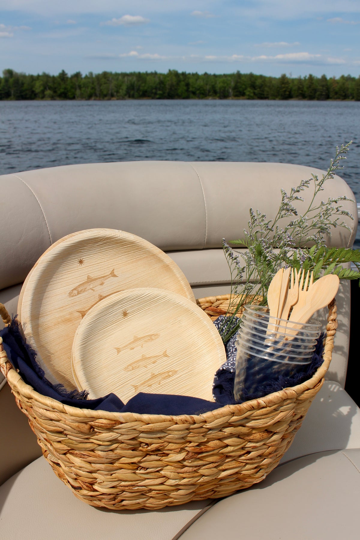 maaterra compostable palm leaf plates with stacked fish design in a basket with sustainable cups and bamboo utensils pictured with the ocean behind. 