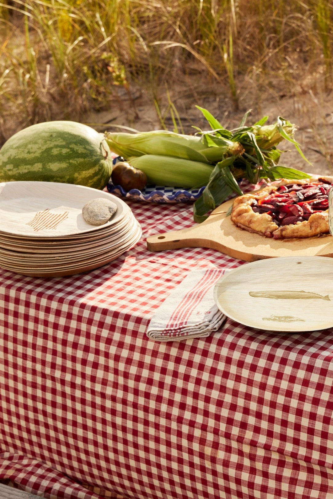 Outdoor setting with a picnic table covered by a red and white checkered tablecloth, featuring maaterra compostable palm leaf plates. 